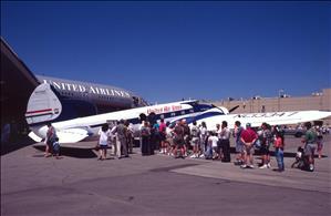 People stand in line next to a small airplane which is next to a large airplane, parked at the airport on a sunny day